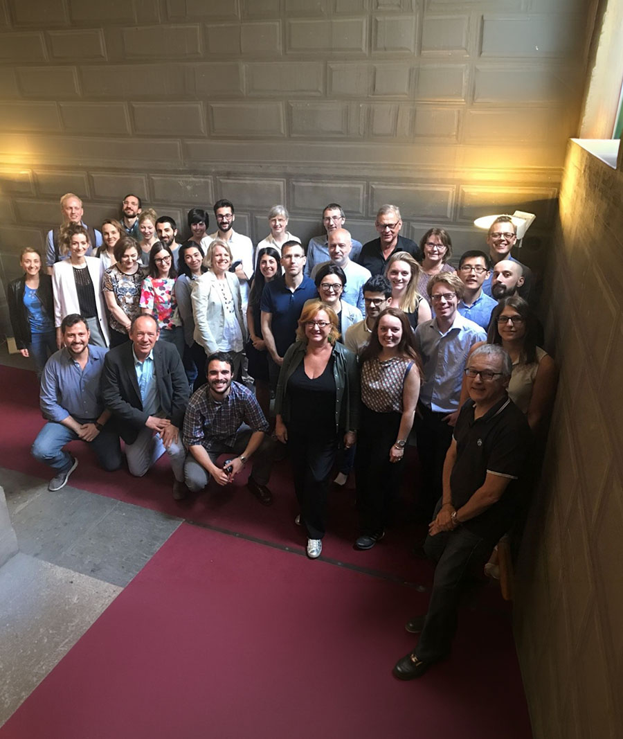 A big group of people pose for a photo in a dimly lit room with a burgundy carpet and grey stone floor.